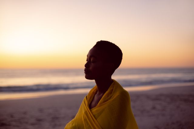 Young woman wrapped in yellow scarf standing on beach with eyes closed during sunset. Ideal for concepts of mindfulness, relaxation, meditation, and tranquility. Suitable for wellness, spiritual, and nature-related content.