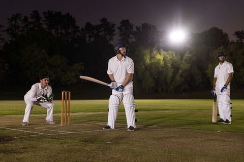 Night cricket match under floodlights with batsman, wicketkeeper and fielders in white