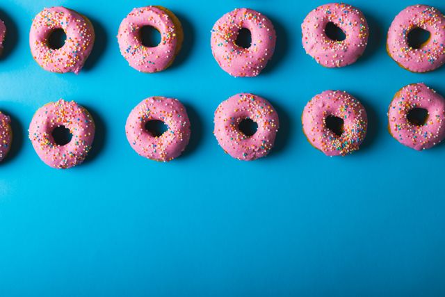 Directly above view of fresh pink donuts arranged side by side with copy space over blue background. unaltered, unhealthy eating and sweet food concept.
