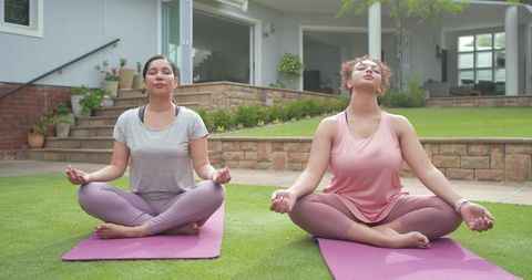 Hispanic and african american women meditating on backyard lawn wearing smartwatches