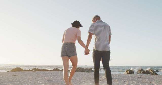Happy diverse couple holding hands and walking on sunny beach by the sea. Summer, vacations, romance, relationship and free time, unaltered.