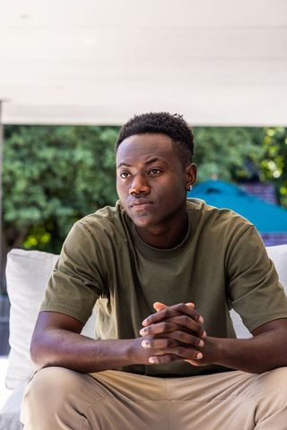 Young african american man sitting contemplative on patio sofa clasping hands, backyard serenity