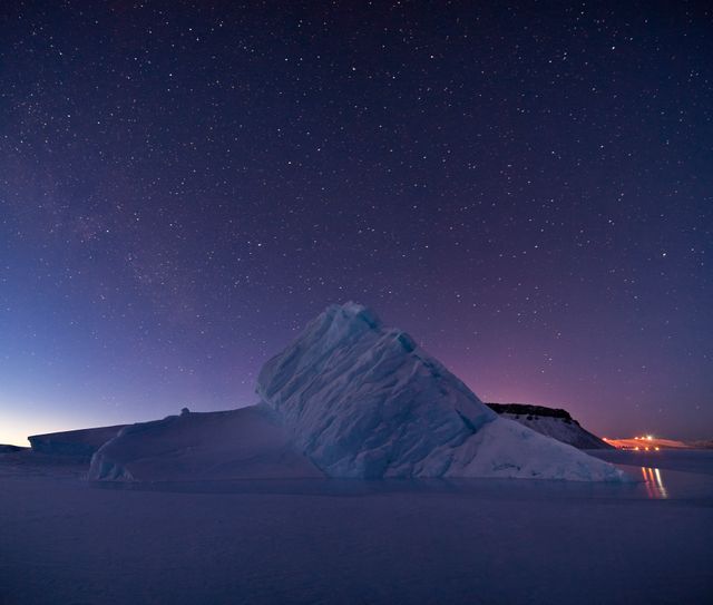Captures serene scene of an iceberg beneath a star-lit sky during twilight. Ideal for themes related to nature, exploration, wilderness, climate change, or celestial photography backdrops.