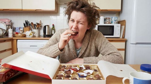 Disheveled woman wincing while eating chocolate from open box on messy kitchen table
