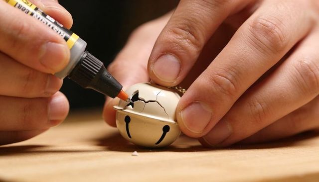 Hands repairing cracked jingle bell using precision glue on wooden workbench