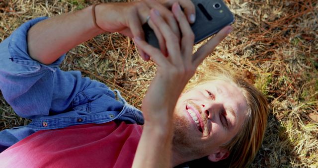 Young man lying on ground outdoors, taking selfie with smartphone, smiling. Casual and relaxed atmosphere surrounded by nature. Perfect for themes like technology, leisure activities, casual lifestyle, youth culture.