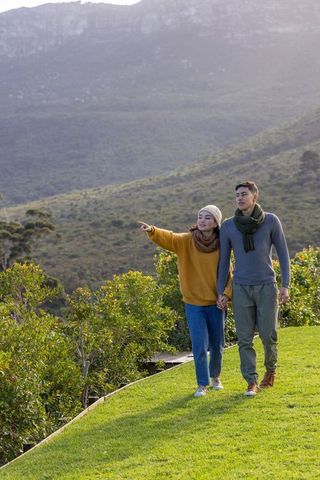 Asian couple walking hand-in-hand on grassy ridge pointing toward distant mountain vista