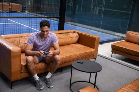 Athlete relaxing courtside checking smartphone on leather sofa in paddle court lounge