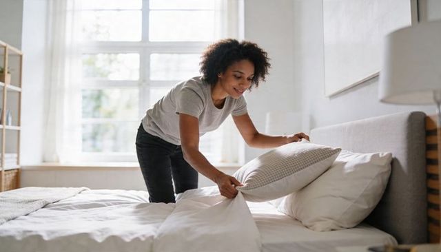 Woman making bed in sunlit minimal bedroom arranging decorative pillow for cozy morning