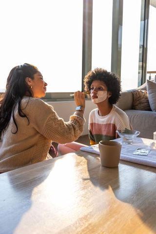 Diverse caregiver applying facial mask on child during cozy warm at-home skincare bonding