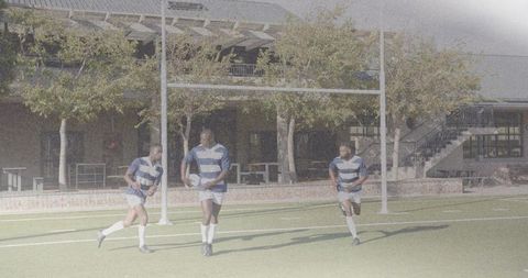Three rugby players running on school turf wearing striped jerseys and carrying ball