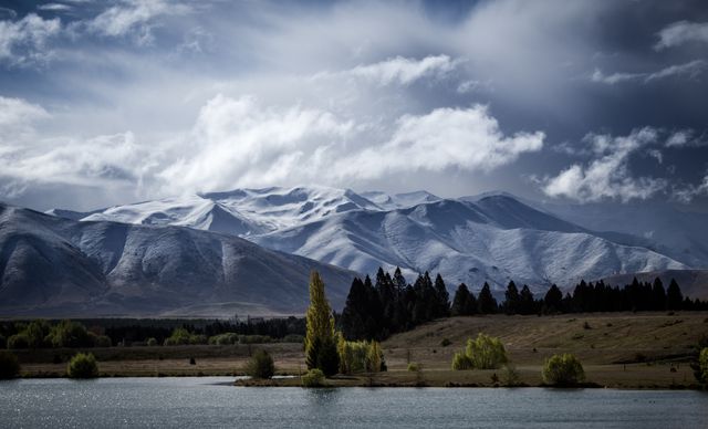 Image features majestic snow-capped mountains set against a dramatic cloudy sky. A tranquil lake and forested area provide a peaceful foreground, creating a striking natural scene. Ideal for use in travel brochures, nature blogs, or environmental campaigns.