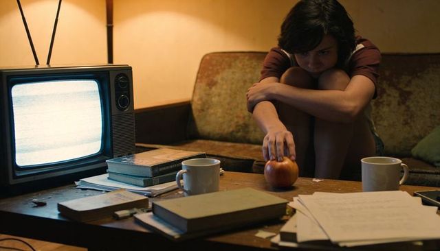 Pensive woman hugging knees reaching for apple on cluttered retro living room table