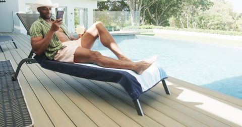Sunlit poolside lounging man wearing straw hat, open green shirt, holding drink and phone