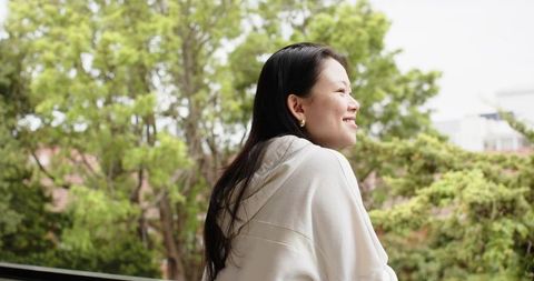 Serene asian woman leaning on balcony railing wearing hoodie smiling toward green trees