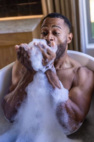 Muscular african american man enjoying bubbly bath, holding foam near face, relaxing in tub