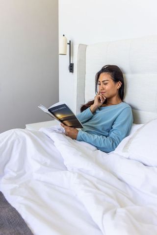 Asian woman reading in cozy minimalist sunlit bedroom, relaxing on bed with paperback