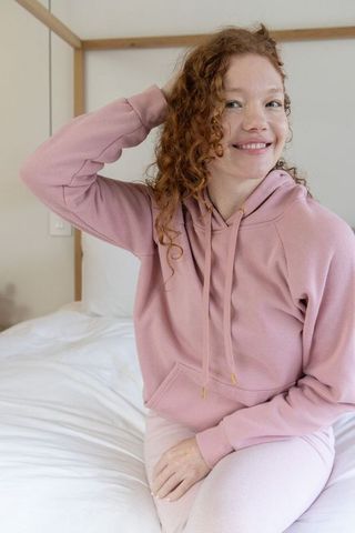 Smiling woman relaxing on bed in pink hoodie and leggings, cozy minimal bedroom morning