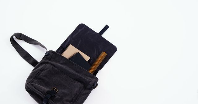 A black shoulder bag with a notebook and pen peeking out lies against a white background, with copy space. Ideal for showcasing everyday carry items or promoting minimalist travel essentials.