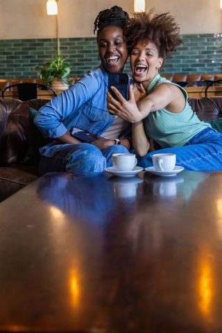 Two african american women laughing and taking selfie on leather sofa in café