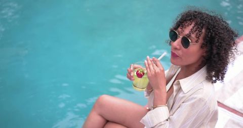 African american woman sitting poolside sipping cocktail wearing sunglasses and shirt