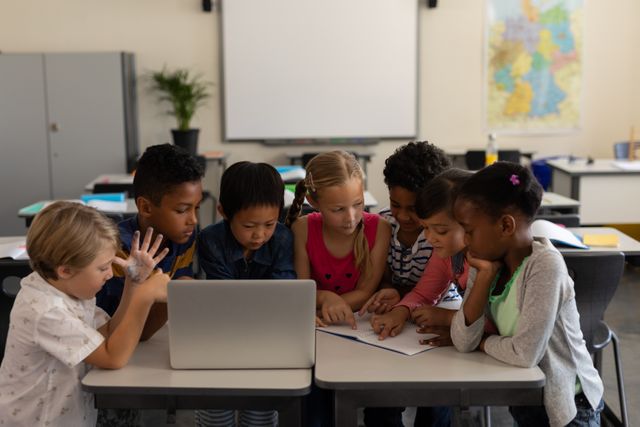 This image shows a diverse group of elementary school children gathered around a laptop and book, engaging in collaborative learning. Ideal for educational content, school websites, and materials promoting teamwork and diversity in education.
