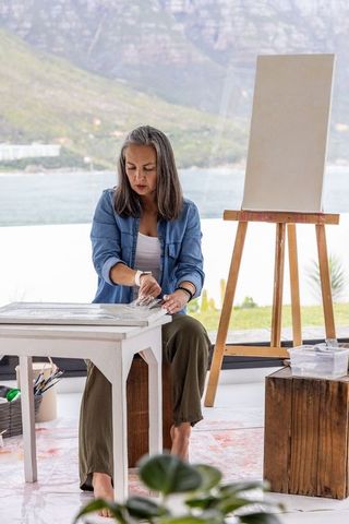 Mature woman creating canvas art in bright waterfront studio with easel and supplies