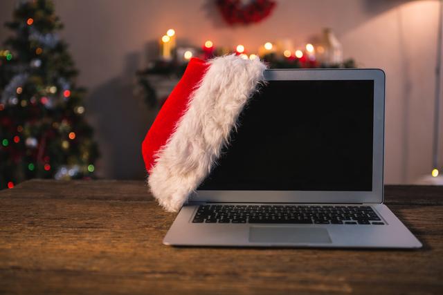 Santa hat hanging on laptop in a cozy home with Christmas decorations and lights. Ideal for holiday-themed marketing, remote work promotions, festive greetings, and technology-related holiday content.