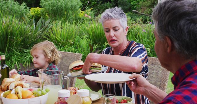Family enjoying an outdoor meal with homemade burgers in a lush garden. Senior woman taking a burger while child happily eats fruit. Perfect for depicting family bonding, summer fun, or homemade food in advertising and marketing materials.