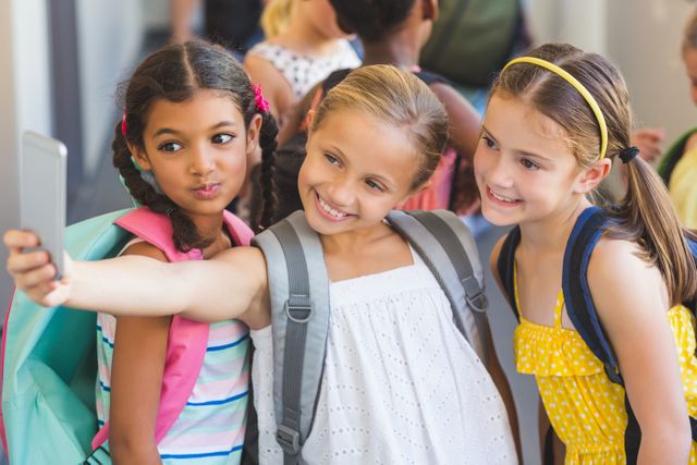 Three school kids are taking a selfie with a mobile phone in a school corridor. They are wearing backpacks and casual clothing, smiling and enjoying their time together. This image can be used for educational content, back-to-school promotions, or articles on childhood friendships and technology in education.