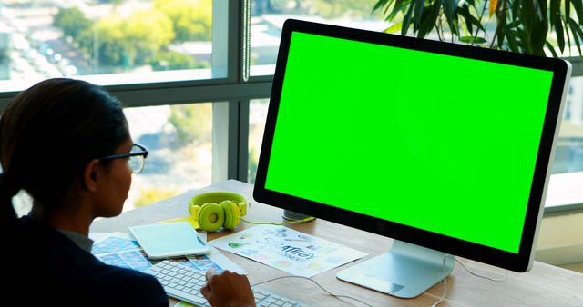 Professional woman working on green screen computer at office desk. Suitable for business, technology, and office workspace themes. Ideal for designs requiring customizable screen content.