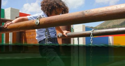 Curly-haired boy climbing wooden rails at outdoor playground wearing safety harness