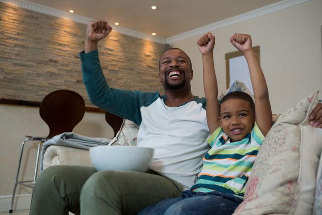 Father and son sitting on couch, raising arms in excitement while watching football match on TV. Perfect for depicting family bonding, sports enthusiasm, and joyful moments at home. Ideal for use in advertisements, blogs, and articles related to family life, sports events, and home entertainment.