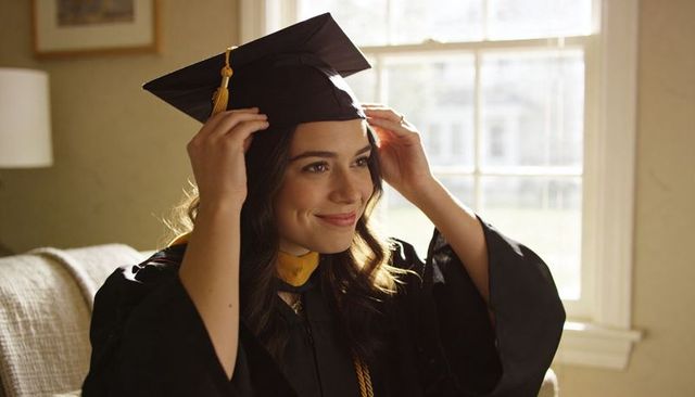 Smiling graduate adjusting cap on couch wearing black gown, gold tassel and stole