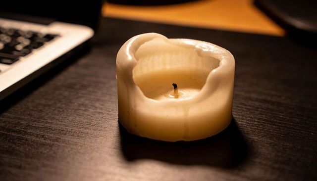 Melted ivory votive candle with charred wick on wooden desk next to laptop under warm lamp