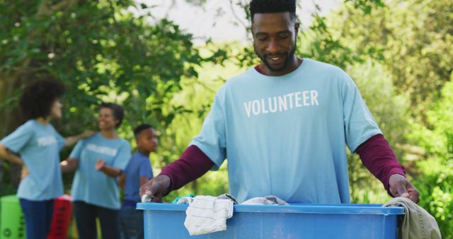 Smiling african american man in volunteer t shirt holding crate, clearing trash outdoors with family. Ecology, volunteering, recycling, nature conservation, family and togetherness.