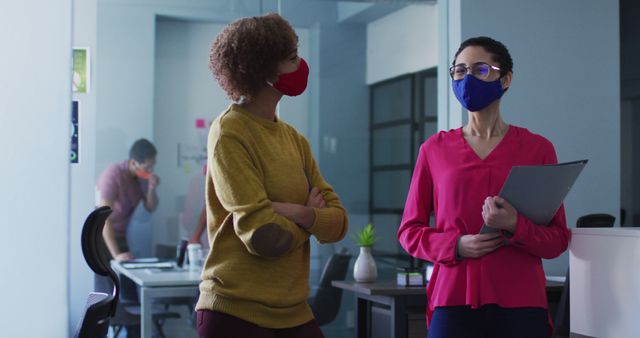 Professional women in office setting practicing safety measures by wearing masks and maintaining distance. This image is ideal for illustrating workplace safety during a pandemic, communication between colleagues, and social distancing practices.