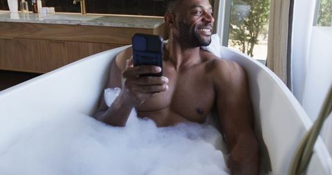 Smiling man relaxing in bubble bath holding flip phone by sunlit window in luxury bathroom