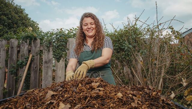 Smiling home gardener turning mulch pile in backyard wearing green apron and gloves