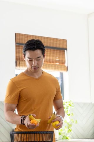 Asian man placing bananas and citrus into basket in bright home kitchen