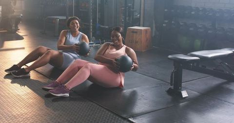Two black women doing partner core training with medicine balls in industrial gym studio