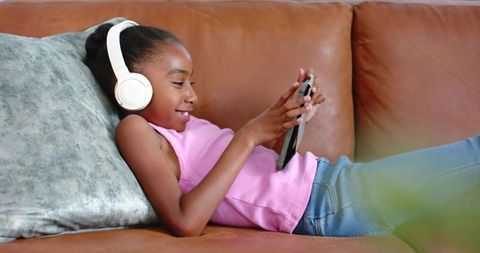African american girl reclining on leather couch listening with white headphones using tablet