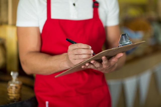 Bakery staff member wearing red apron writing on clipboard at counter in bake shop. Useful for illustrating concepts related to small business operations, inventory management, retail work, and food service industry. Ideal for articles, blogs, and marketing materials focusing on bakery business, employee tasks, and organizational skills.