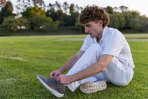 Young man tying shoelace on grass in white sportswear during golden hour practice