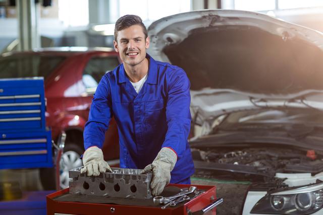 Smiling mechanic in blue uniform checking car parts in a repair garage. Ideal for use in automotive service promotions, mechanic training materials, auto repair service advertisements, and content related to vehicle maintenance and repair.