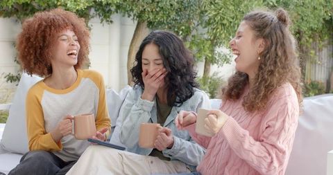 Diverse women laughing and sipping coffee on outdoor sofa, casual backyard gathering