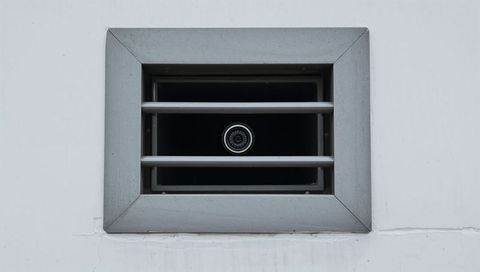Security camera peering through rectangular metal vent with horizontal grille