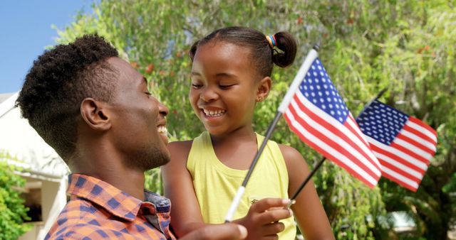 Father and daughter smiling and holding American flags in park. Ideal for representations of patriotism, family bonding, Independence Day celebrations, and outdoor summer activities. Suitable for use in promotional materials, advertisements, and articles focusing on family events and festive occasions.