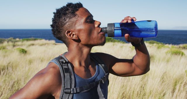 Young man in athletic attire drinking from water bottle while hiking near ocean. Scenic outdoor activity on sunny day emphasizes hydration, fitness, and enjoyment of nature. Ideal for promoting outdoor adventures, healthy lifestyles, and hiking gear.