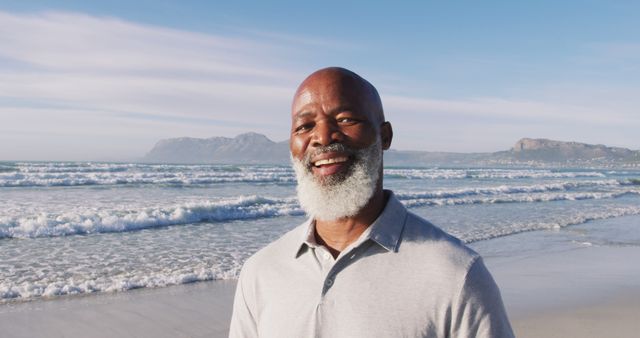 Senior man with grey beard smiling at the beach with ocean waves in the background. Ideal for use in advertisements promoting a healthy and joyful lifestyle, retirement plans, travel experiences, and wellness activities. Suitable for articles and blog posts related to outdoor adventures, vacation planning, and personal stories of enjoyment and fulfillment.